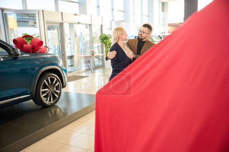 Téléchargez les photos : Mari et femme se tiennent près de leur voiture donnée enveloppée dans une couverture rouge. - en image libre de droit