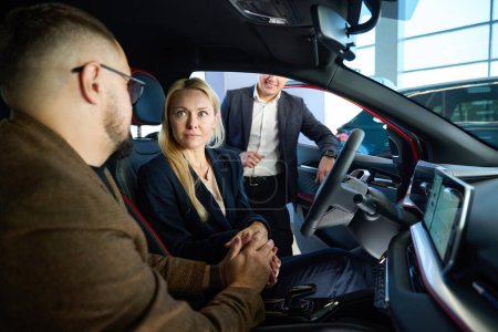 Téléchargez les photos : Jeune vendeur de voitures montre l'intérieur d'une nouvelle voiture à un jeune couple caucasien - en image libre de droit