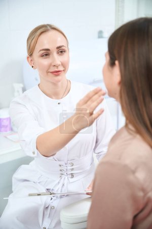 Photo for Professional cosmetologist sitting in hospital, looking at patient and checking skin of lady - Royalty Free Image