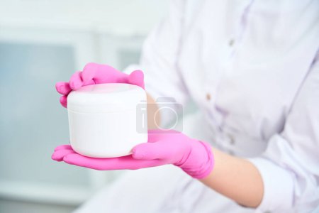 Téléchargez les photos : Photo en gros plan de la femelle en gants roses tenant le flacon avec le produit de soins de la peau et le présentant - en image libre de droit