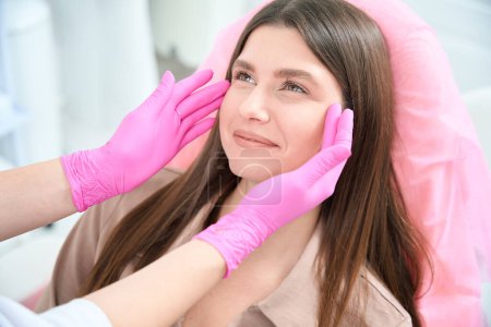 Photo for Smiling lady looking at camera, cosmetologist in pink gloves touching face of client - Royalty Free Image