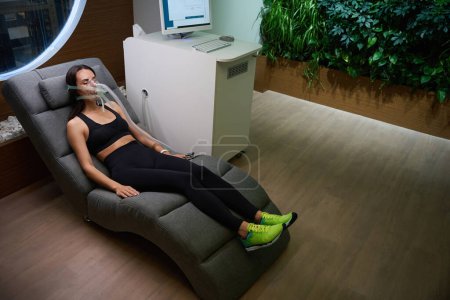 Young female in sporty clothes lying on comfort chair in mask, undergoing hypoxic therapy
