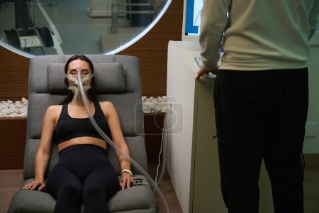Dame en vêtements sportifs couchée sur une chaise confort en masque, subissant une thérapie hypoxique