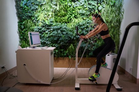 Young lady in sporty clothes in mask riding bike, undergoing Spiro cycle ergometry in gym