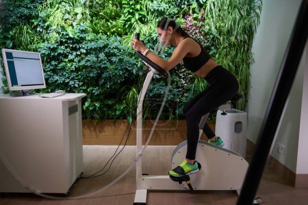 Sporty lady in sporty clothes in mask riding bike, undergoing Spiro cycle ergometry in gym