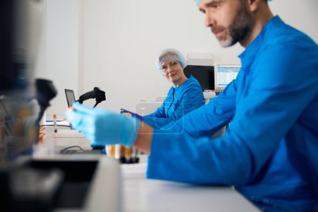 Téléchargez les photos : Homme virologiste travaille avec des échantillons de sang, sa collègue féminine travaille à proximité - en image libre de droit