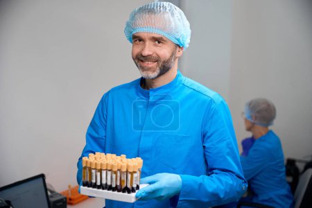 Asistente de laboratorio masculino sostiene en sus manos un conjunto con biomaterial para un análisis de sangre, las muestras están marcadas
