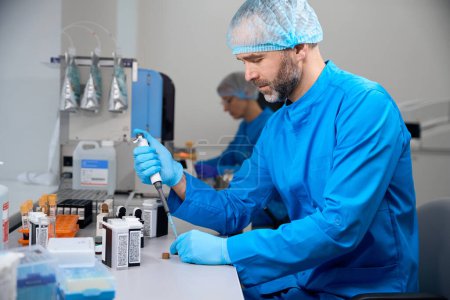 Foto de Asistente de laboratorio masculino examina material biológico en un laboratorio moderno, su colega trabaja cerca - Imagen libre de derechos