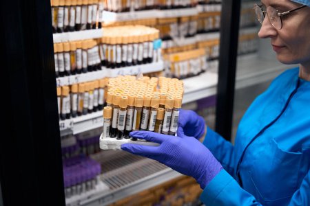 Téléchargez les photos : Assistante de laboratoire féminine tient dans ses mains un ensemble avec biomatériau pour l'analyse de sang, les échantillons sont stockés dans le réfrigérateur - en image libre de droit