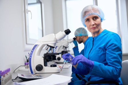 Frau mittleren Alters und ein männlicher Kollege arbeiten in einem modernen Labor, ein leistungsstarkes Mikroskop kommt zum Einsatz