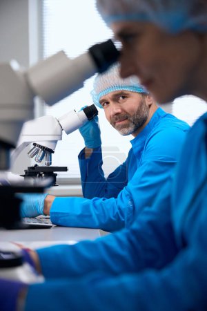 Mann mittleren Alters und eine Kollegin arbeiten in einem modernen Labor, ein leistungsstarkes Mikroskop kommt zum Einsatz