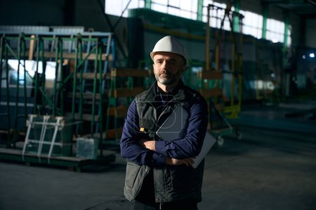 Photo for Middle-aged man stands in a production workshop, he is wearing a safety helmet - Royalty Free Image