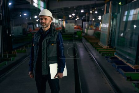 Engineer with a folder stands in a production workshop, a man in a safety helmet