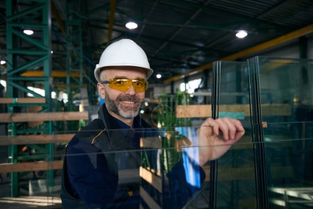 Foto de El hombre con gafas de seguridad y un sombrero duro trabaja en un taller de producción, las precauciones de seguridad se observan en la producción de ventanas - Imagen libre de derechos