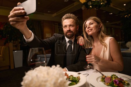 Téléchargez les photos : Belle femme avec son compagnon prendre un selfie dans un restaurant, une dame dans une robe de soirée - en image libre de droit