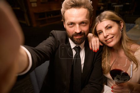 Téléchargez les photos : Couple élégant prenant un selfie dans un restaurant, dame avec un verre de vin rouge - en image libre de droit