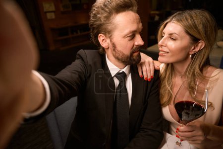 Téléchargez les photos : Elégant couple d'âge moyen prend un selfie dans un restaurant, la dame avec un verre de vin rouge - en image libre de droit