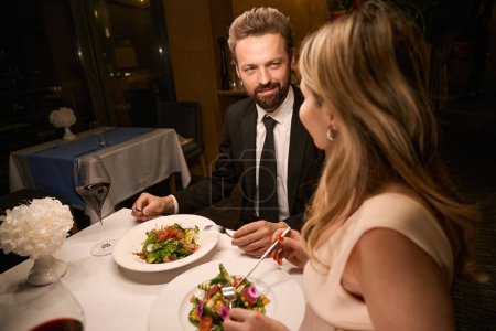 Foto de Hombre y mujer disfrutando de una cena romántica en un acogedor restaurante, ensalada ligera y vino servido - Imagen libre de derechos