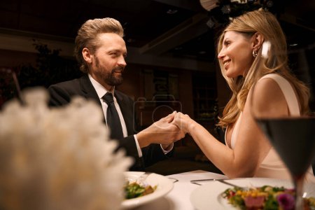 Foto de Hombre y mujer de mediana edad se toman de la mano y se miran, la pareja se sienta en una mesa en un restaurante - Imagen libre de derechos