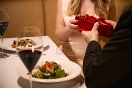 Foto de Mujer aceptando una caja roja con un regalo de su acompañante, pareja cenando en un acogedor restaurante - Imagen libre de derechos