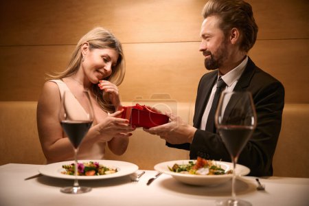 Lady accepts a gift in a red box from a man, a couple has dinner in a cozy restaurant