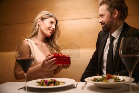 Foto de Señora recibió un regalo de un hombre en una caja roja, la pareja está cenando en un acogedor restaurante - Imagen libre de derechos