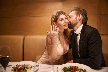 Glückliche Frau zeigt einen Ring an ihrer Hand, ihr Begleiter küsst sie zärtlich