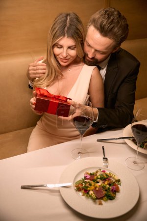 Téléchargez les photos : Homme arrangé une magnifique surprise pour son charmant compagnon lors d'un dîner romantique, un cadeau dans une boîte rouge - en image libre de droit