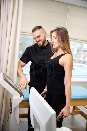 Physiotherapist adjusting diagnostic machine for middle aged woman, female patient in comfortable clothes