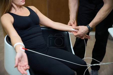 Photo for Specialist attaches electrodes to a woman wrists for a diagnostic procedure, people are in a wellness center - Royalty Free Image