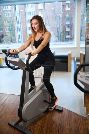 Photo for Female exercises on an exercise bike in a health center, she is in a comfortable training suit - Royalty Free Image