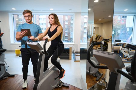 Téléchargez les photos : Instructeur dans une salle de gym surveille une séance d'entraînement des clients sur un vélo d'appartement, il ya beaucoup de machines d'exercice dans la salle - en image libre de droit