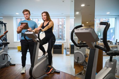 Téléchargez les photos : Trainer contrôle la formation des clients sur un vélo d'appartement, il y a beaucoup de machines d'exercice dans la salle - en image libre de droit