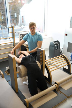 Woman in gym works on stretching her muscles under supervision of a physiotherapist, she is wearing a black workout suit