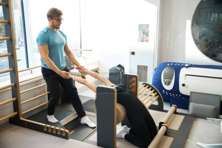 Photo for Female working on stretching muscles under the supervision of a physiotherapist, using a special exercise machine - Royalty Free Image