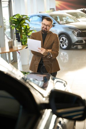 Téléchargez les photos : Jeune homme en lunettes regarde un contrat pour l'achat d'une nouvelle voiture - en image libre de droit