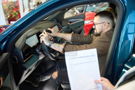 Téléchargez les photos : Homme à lunettes et un costume tient le volant d'une nouvelle voiture et l'évalue - en image libre de droit