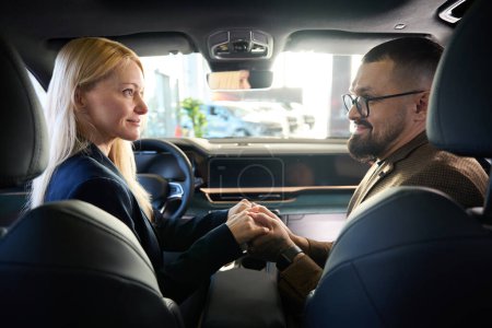 Téléchargez les photos : Femme tient les mains de son mari assis à l'intérieur d'une nouvelle voiture dans une salle d'exposition de voiture - en image libre de droit