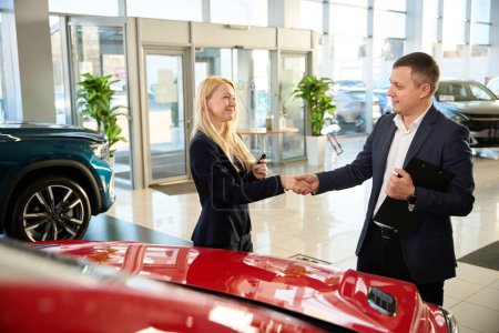 Téléchargez les photos : Consultant automobile serrant la main d'une femme à côté d'une nouvelle voiture rouge - en image libre de droit