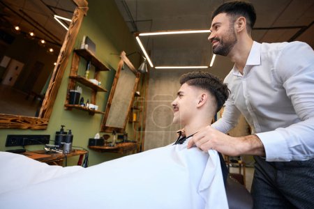 Téléchargez les photos : Homme assis dans un fauteuil et coiffeur professionnel le couvrant de robe de coiffure dans le salon de coiffure - en image libre de droit