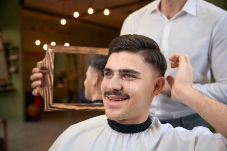 Téléchargez les photos : Coiffeur tenant miroir et montrant le résultat de son travail à l'homme dans le salon de coiffure - en image libre de droit