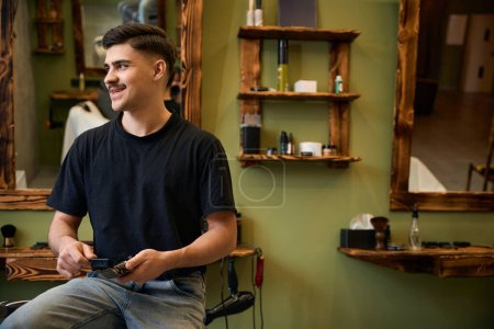 Foto de Peluquería masculina sonriente limpiando el cortador de cabello con cepillo mirando hacia otro lado en la barbería - Imagen libre de derechos