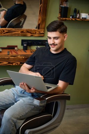 Téléchargez les photos : Barber communiquer en ligne via ordinateur portable assis dans la chaise de coiffure - en image libre de droit