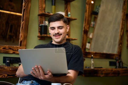 Téléchargez les photos : Coiffeur homme souriant utilisant un ordinateur portable pour le travail naviguant nouvelles idées de coiffure au lieu de travail de salon de coiffure - en image libre de droit