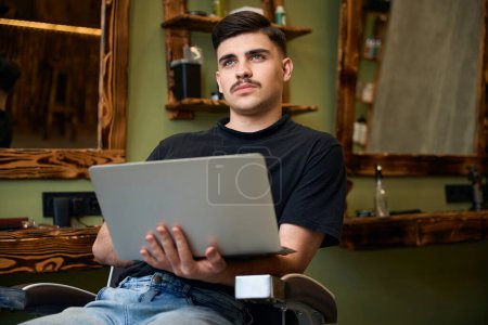 Téléchargez les photos : Homme réfléchi barbier à l'aide d'un ordinateur portable pour la navigation de nouvelles idées de coiffure au lieu de travail de salon de coiffure - en image libre de droit