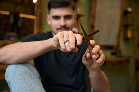 Photo for Handsome barber showing scissors while sitting at his workplace in barbershop - Royalty Free Image