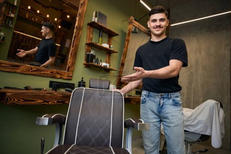 Téléchargez les photos : Barbier professionnel debout sur son lieu de travail invitant les clients dans le salon de coiffure - en image libre de droit