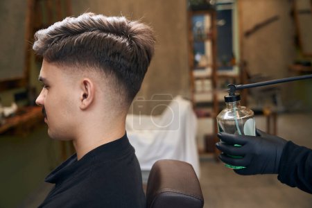 Man barber sprinkles perfume from vintage designed stylish bottle above clients hair in barbershop