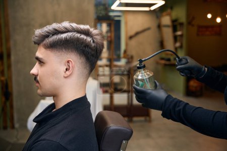 Foto de Peluquería irreconocible rociando perfume en el hombre barbudo en la capa de peluquería en la barbería - Imagen libre de derechos