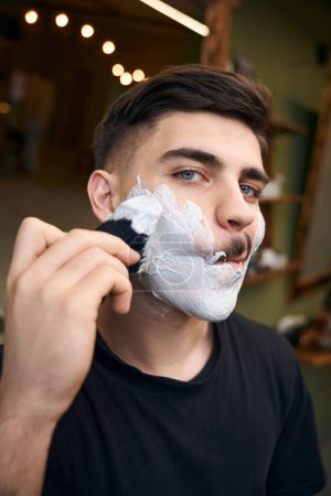 Foto de Joven con espuma en la cara afeitándose en la barbería - Imagen libre de derechos